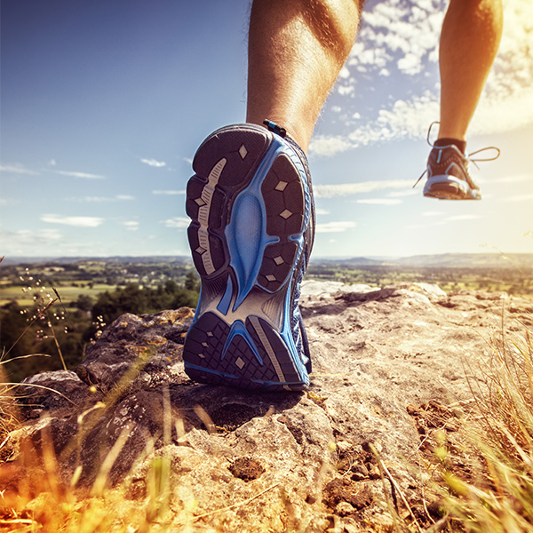 The image shows a person s foot with a blue and black hiking shoe on it, placed in the foreground against a backdrop of a rocky terrain under a clear sky.