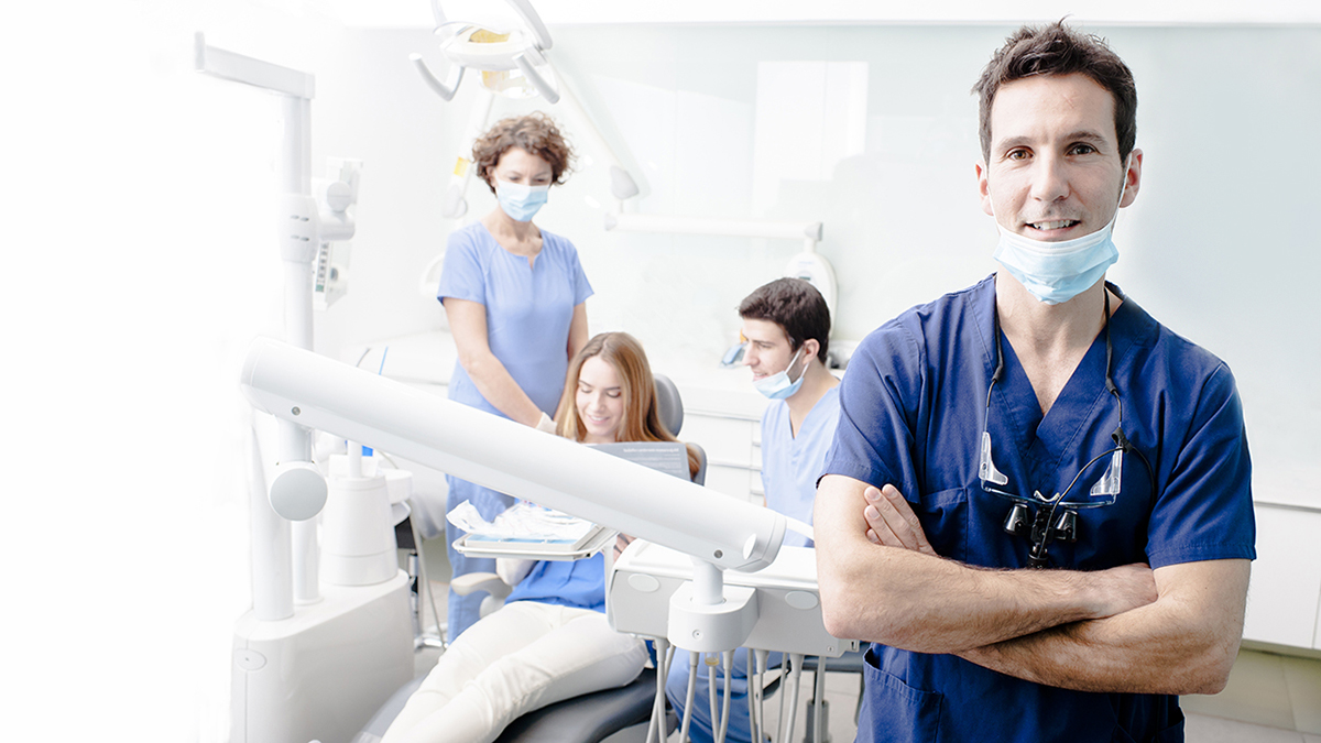 The image features a man in scrubs standing confidently in front of a dental chair, with a group of dental professionals and patients behind him, all set within a modern dental clinic.