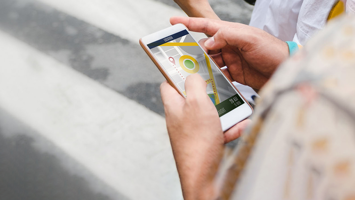 The image shows a person holding a smartphone with a map displayed on the screen, taken from an angle that includes part of the person s body and the urban street environment.