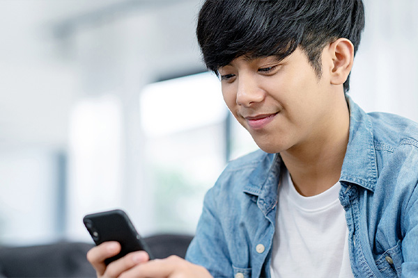 A young man in a blue denim shirt is holding a smartphone and smiling at the camera.