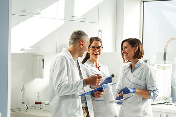 This is a color photograph featuring three individuals in what appears to be a laboratory setting, with two people wearing white lab coats and one person in a blue shirt. They are standing near each other, engaged in conversation or activity, with various pieces of equipment and scientific materials visible around them.