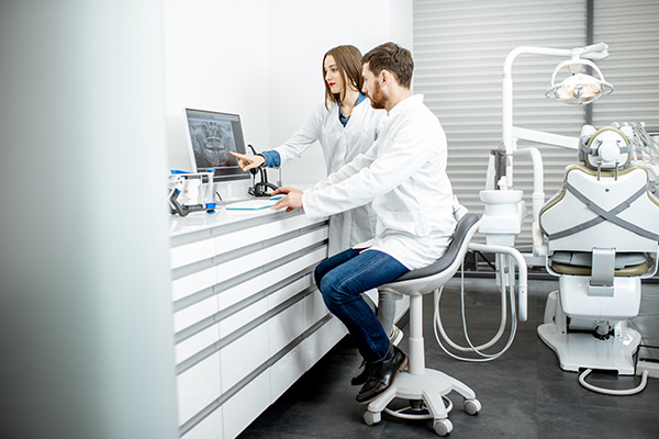 A man and woman in a medical or dental office, with the man standing at a desk with a computer monitor and the woman seated on a stool; they are both dressed in white lab coats.