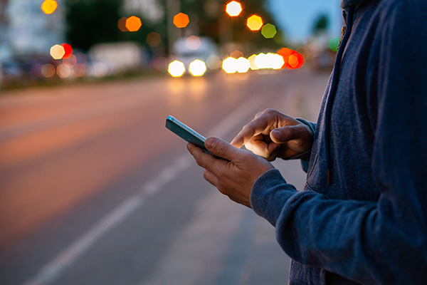 An adult is standing on a sidewalk at night, looking at a smartphone.