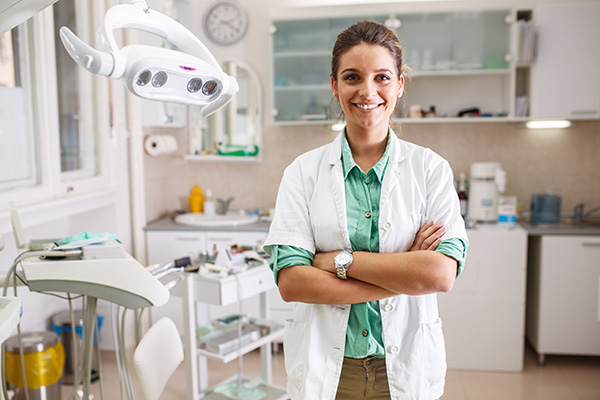 A woman in a white coat stands confidently in front of dental equipment, radiating professionalism.