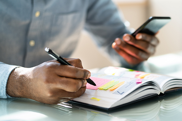 A person is sitting at a desk, holding a smartphone in their hand and looking at it, while also writing or taking notes on an open book that is placed on the table.