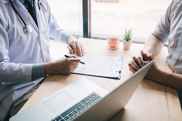 A medical professional in a white coat is seated at a table with a laptop, reviewing patient information and engaging in conversation with another individual.