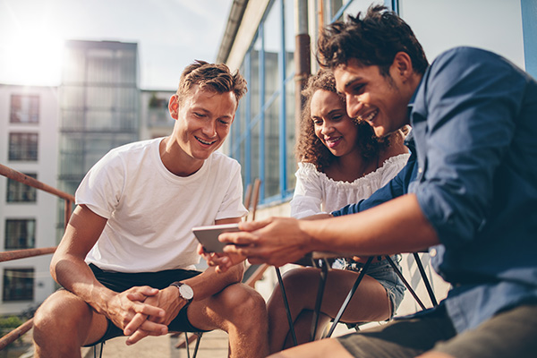 The image depicts three individuals engaged in a conversation while seated outdoors, with a tablet visible between them.