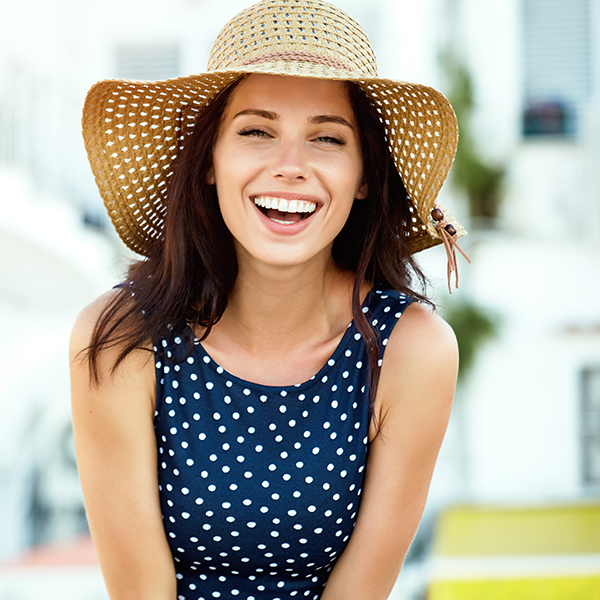 The image shows a woman wearing a straw hat, smiling broadly at the camera, with her hand on her hip. She is dressed in a sleeveless top and has a polka dot pattern on it.