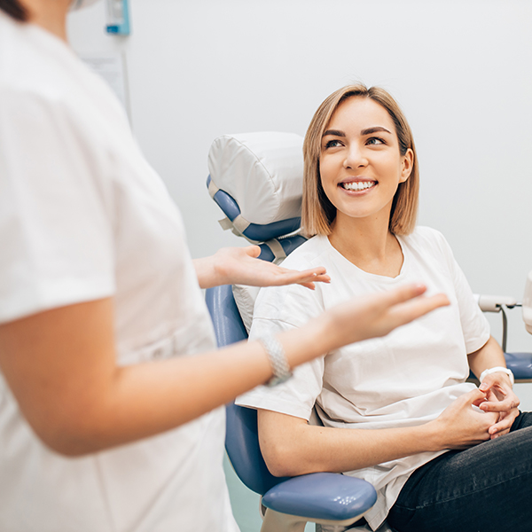 A woman in a white shirt is seated in a dental chair, smiling and engaging with two dental professionals who are standing beside her.