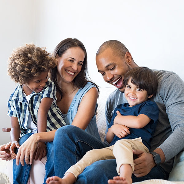 The image shows a family of four, including an adult couple and two young children, sitting together on a couch, smiling and looking at the camera.