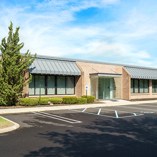 The image shows a modern, one-story brick building with a flat roof and large glass windows. It appears to be an office or commercial building, possibly serving as a headquarters, given its size and architectural style. There is a parking lot in front of the building, with several vehicles parked. A tree with green leaves can be seen on the left side of the image. The sky is partly cloudy, suggesting it might be a pleasant day.