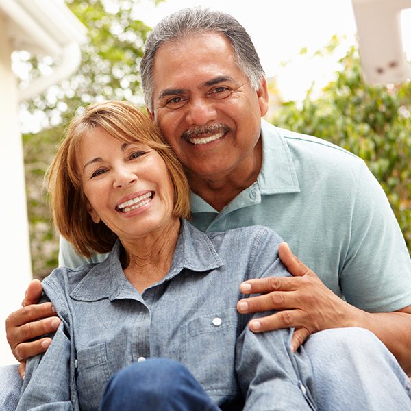 An elderly couple embracing, with the man wearing a blue shirt and the woman in a white top, smiling and posing for a photo.