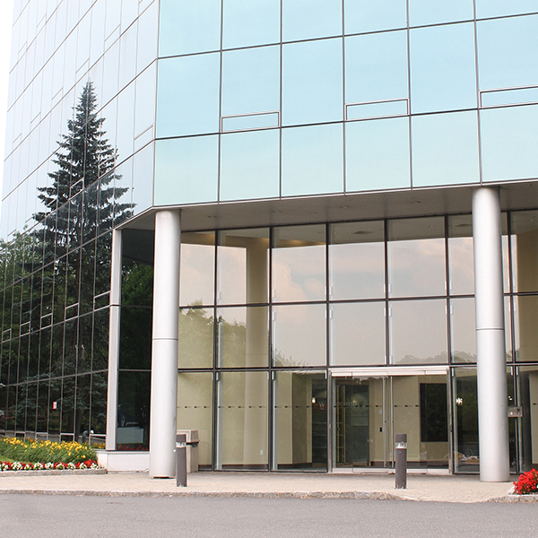An image of a modern building entrance with large glass doors, reflecting the surrounding environment and sky, during daylight hours.