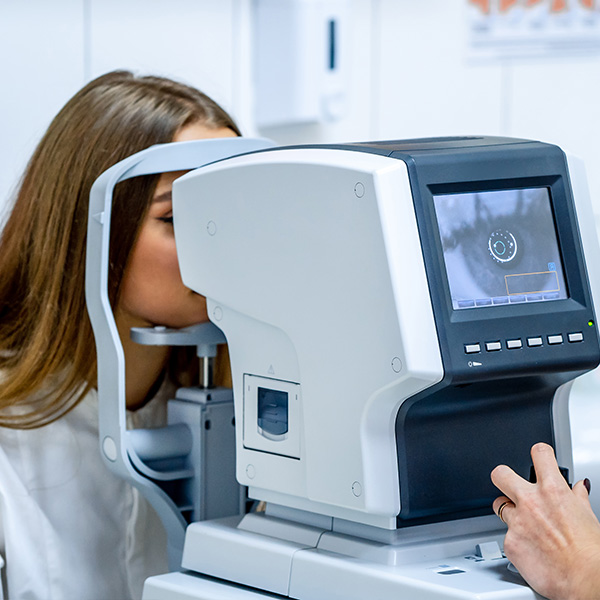 A woman is leaning over a large, modern eye examination machine, with a technician in the background adjusting settings on the device.