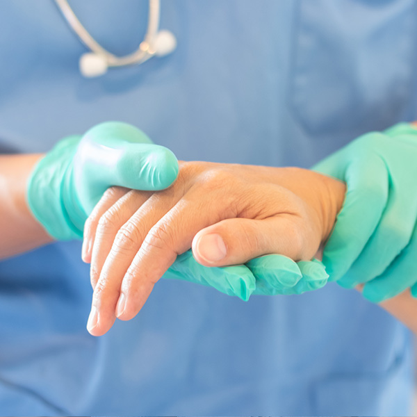 Healthcare professional in blue scrubs, wearing green gloves and a stethoscope, examines a hand with a medical device.