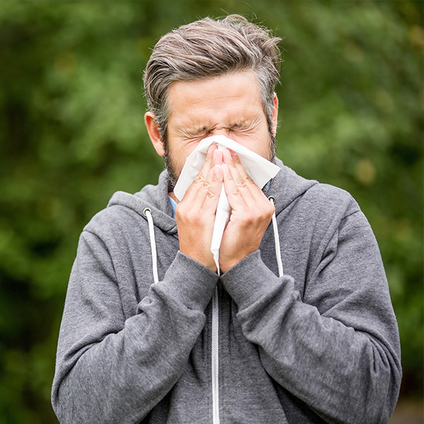 A man with a beard, wearing a gray sweatshirt and a blue mask over his mouth and nose, is wiping his face with a handkerchief.