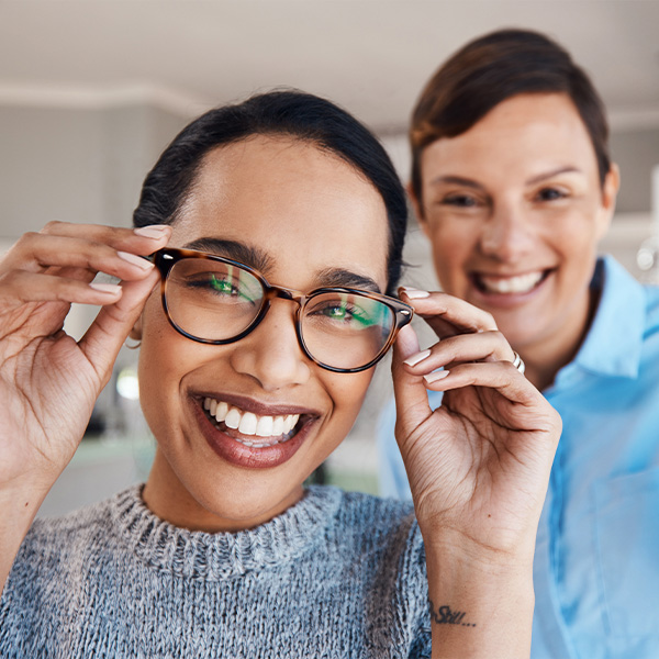 The image features a person wearing glasses and smiling, with another individual standing behind them, both in an indoor setting.