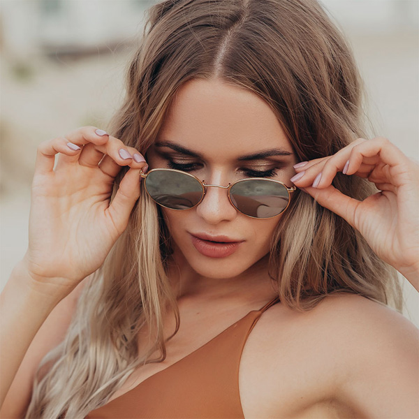 A woman wearing sunglasses and a brown tank top, with her hand on her face, stands in front of a beach setting.