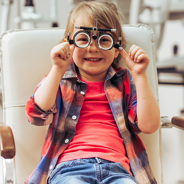 A young girl wearing glasses and a plaid shirt is seated in an eye examination chair, smiling at the camera.