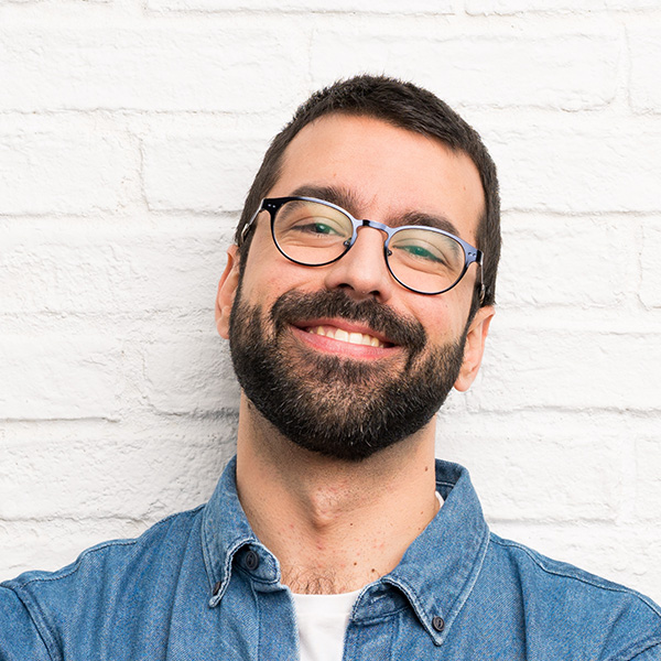 The image features a man with a beard, wearing glasses and a blue denim shirt, smiling at the camera against a white brick wall background.