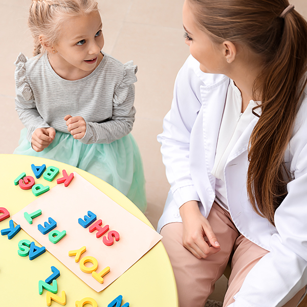 A young girl is sitting at a table with a woman who appears to be a medical professional, both are looking at an alphabet-themed educational activity.
