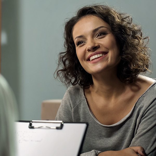 A woman with short hair, smiling and seated in a room, engaged in conversation with another person who is partially visible.