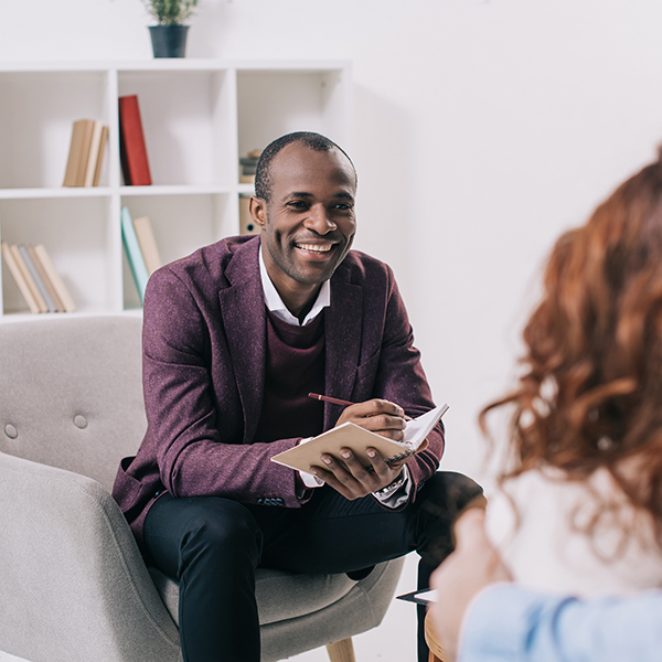 The image shows a man sitting on a couch, smiling at the camera, with a notebook in his hand. He appears to be engaged in a conversation or interview with another person who is not visible in the frame.