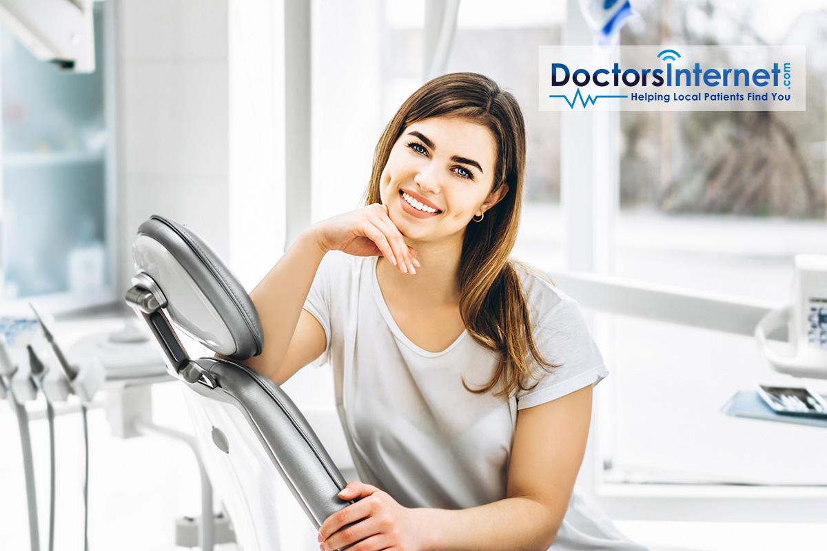 A woman in a dental office, standing by a sink with a mirror above it, smiling and looking directly at the camera.