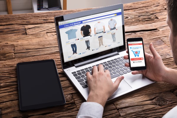 A person using a laptop to browse a fashion e-commerce website, with a smartphone in hand displaying the same site.