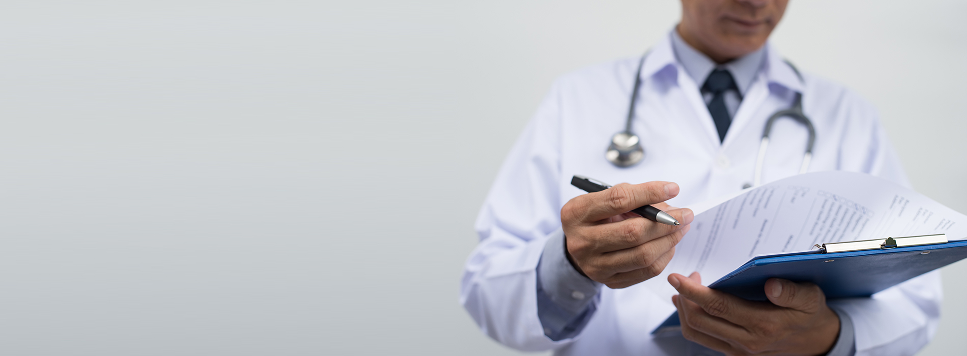 The image shows a male doctor in a white coat, holding a clipboard and pen, writing or reviewing notes while standing against a white background.