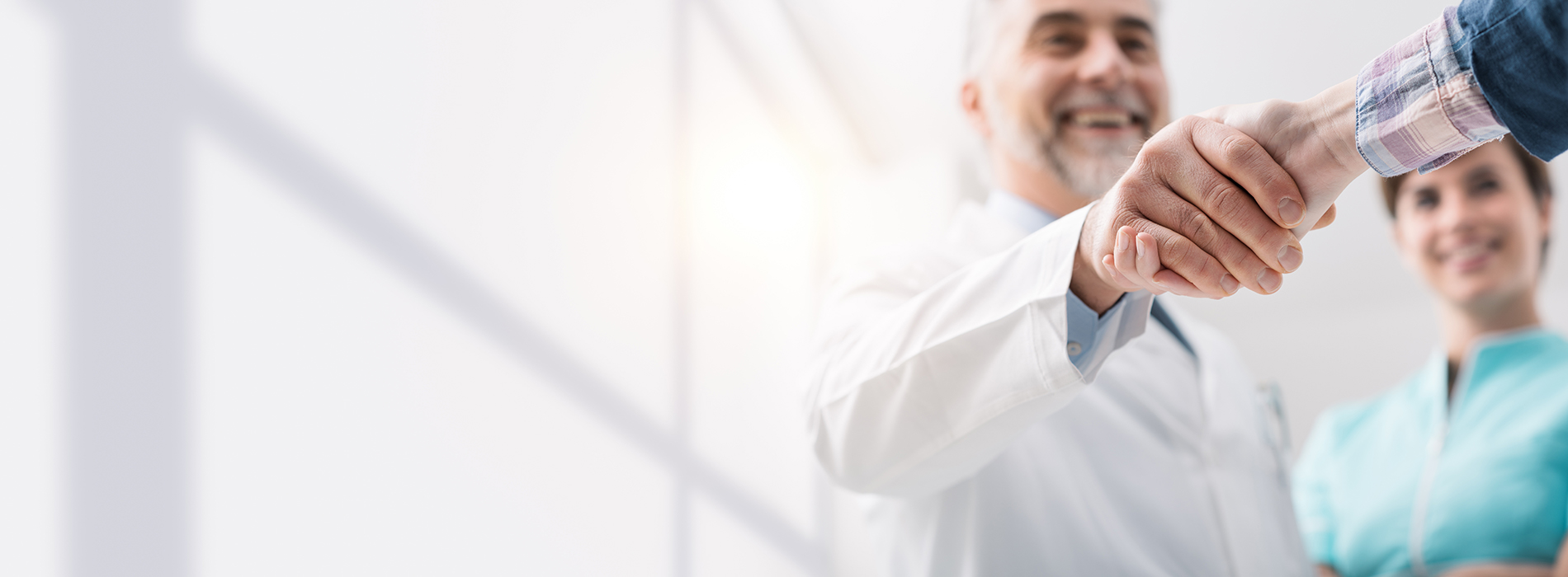 The image shows two individuals shaking hands, with one person holding a smartphone. They are in an indoor setting that appears to be a professional environment, possibly a business or office space, as suggested by the presence of a window with blinds and the attire of the people present.