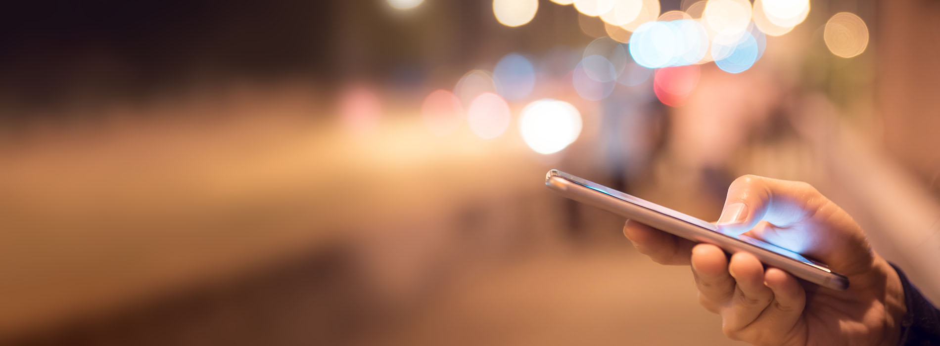 The image shows a person holding a smartphone with their thumb, likely using it to capture a photo or view content. The phone is held up close to the camera, making the screen and the person s hand visible. In the background, there is a blurred street scene with traffic lights and cars, indicating an urban setting at night.