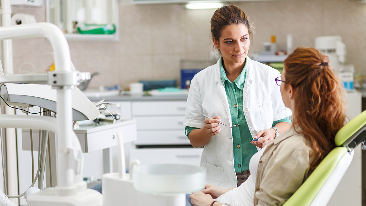 A woman is assisting a patient in a dental office, with the dental chair visible and various dental tools nearby.