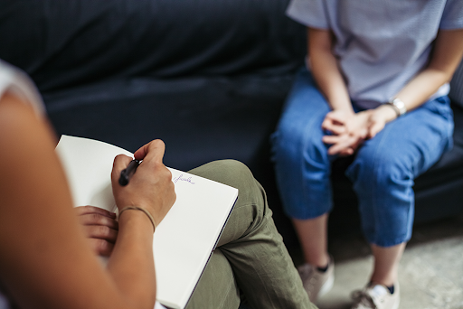 The image shows a person sitting on the floor, holding a clipboard and pen, engaged in conversation with another individual who is seated on a couch.