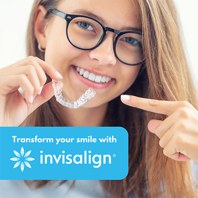 The image shows a young woman with a radiant smile, holding a clear aligner up to her teeth. She is pointing to the aligner with her finger. The background is blurred but suggests an indoor setting, possibly a dental office or a room with a white wall.