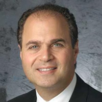 The image shows a man wearing a dark suit, posing with a slight smile against a neutral background.