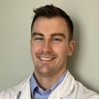 The image features a man wearing a white lab coat, smiling, and posing for the photo. He has short hair and is standing against a plain background.