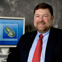 The image shows a man sitting at a desk with a computer monitor, smiling and looking directly at the camera.