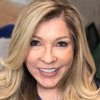 The image features a woman with long blonde hair, wearing makeup and a light blue top. She has a bright smile and appears to be indoors.