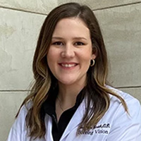 The image shows a woman wearing a white lab coat and smiling at the camera. She has short blonde hair, is standing against a neutral background with a wall-mounted credential to her left, and appears to be posing for a professional portrait.